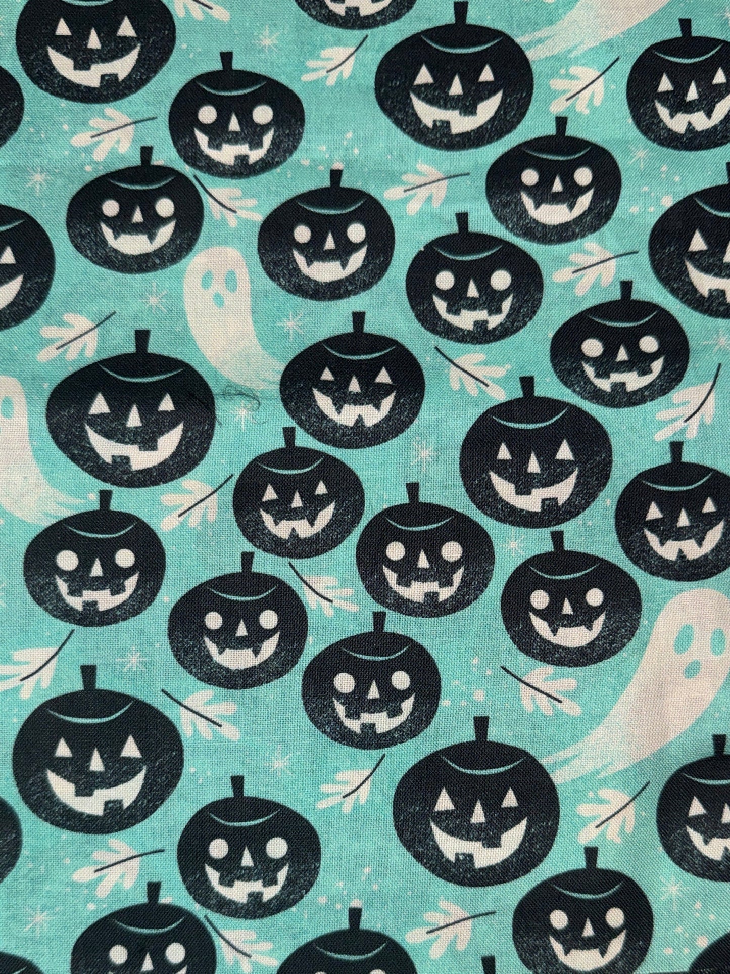A close-up of a Halloween-themed fabric pattern featuring black jack-o'-lanterns, white ghosts, and small bats on a light aqua background.